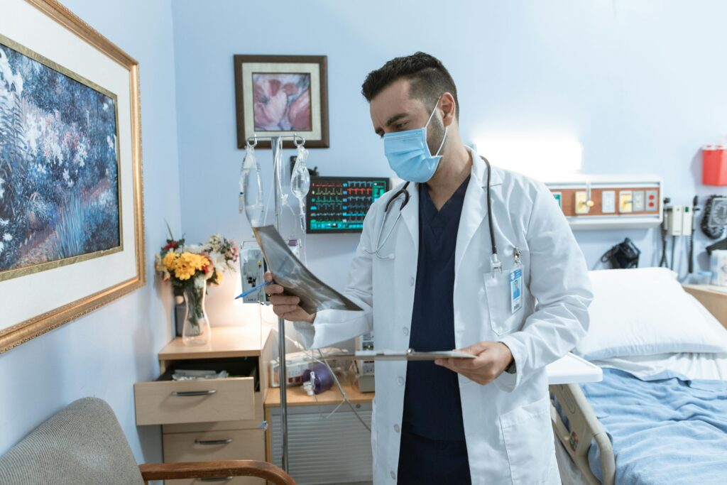pexels-photo-6129049-6129049 A male doctor wearing a face mask reviews an X-ray in a hospital room, focusing on patient diagnosis.